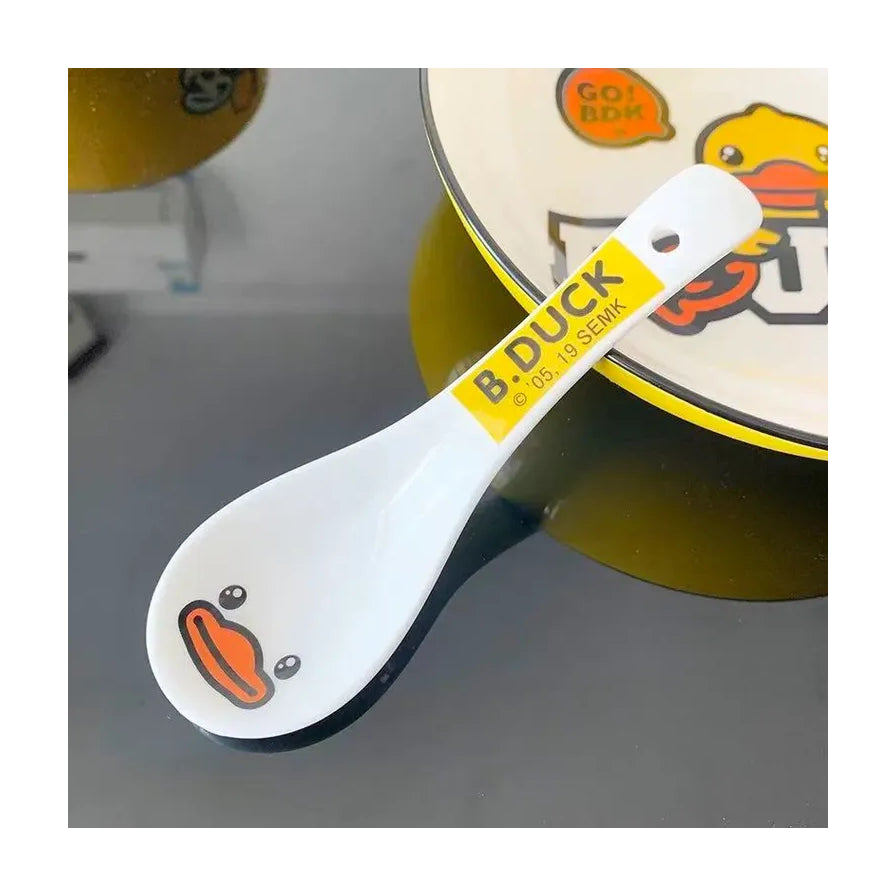 B.DUCK 萌宠小鸭陶瓷汤勺放置在桌面，背景可见同系列餐盘，展示生活化使用场景。