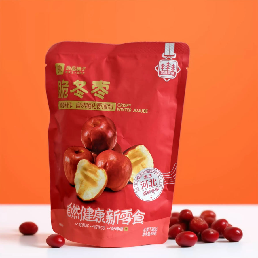 良品铺子脆冬枣包装袋立于白色台面，背景为橙色，周围散落着数颗红润的脆冬枣实物。
