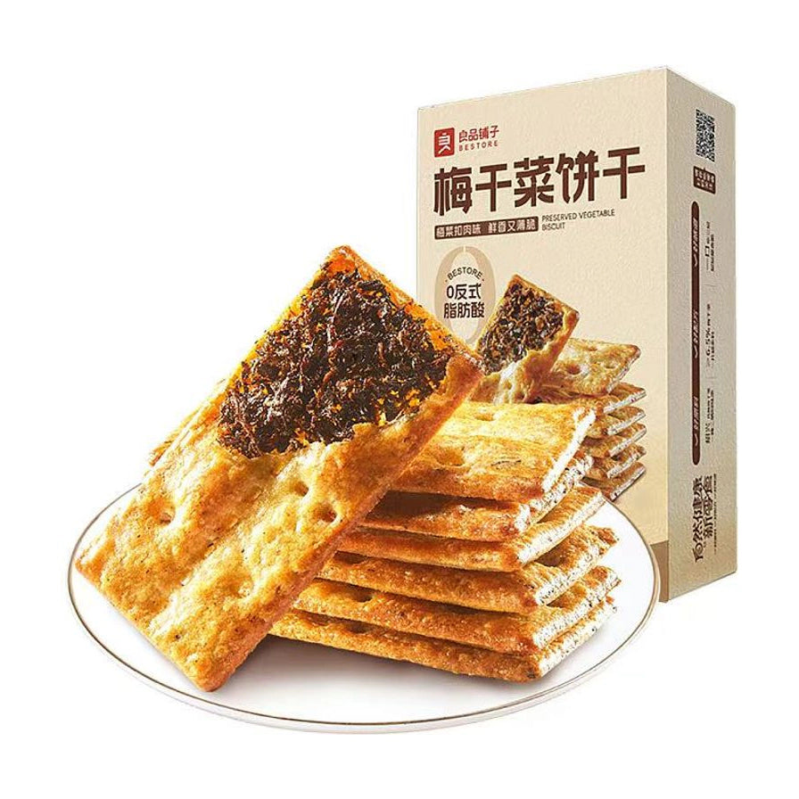 良品铺子梅干菜饼干，白色圆盘上叠放着多片酥脆的梅干菜饼干，背景为商品包装盒，包装上印有0反式脂肪酸字样。