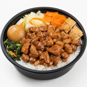 俯拍视角下的大碗桶饭，黑色圆桶内盛满白米饭，铺有酱汁浓郁的肉块、半颗卤蛋、豆腐块、胡萝卜片和青菜。