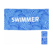 白色背景下展示的蓝色斑马纹沙滩浴巾，印有SWIMMER字样，右下角展示其折叠收纳后的圆柱状形态。