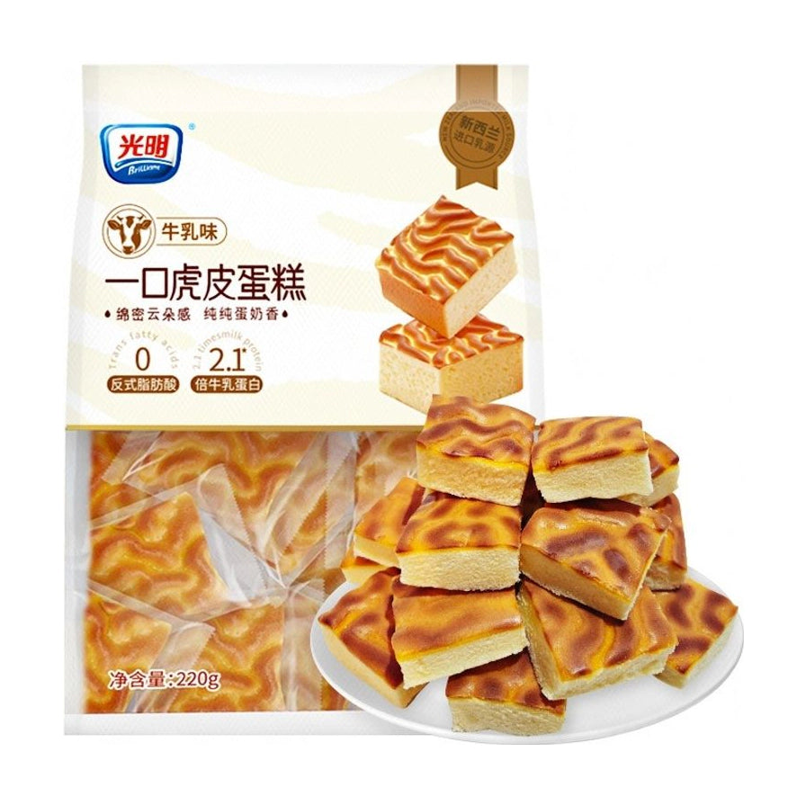 光明一口小虎皮蛋糕牛乳味包装袋，展示了内部独立包装的蛋糕块和一盘切成方块的虎皮蛋糕，包装上标有0反式脂肪酸和2.1倍牛乳蛋白。