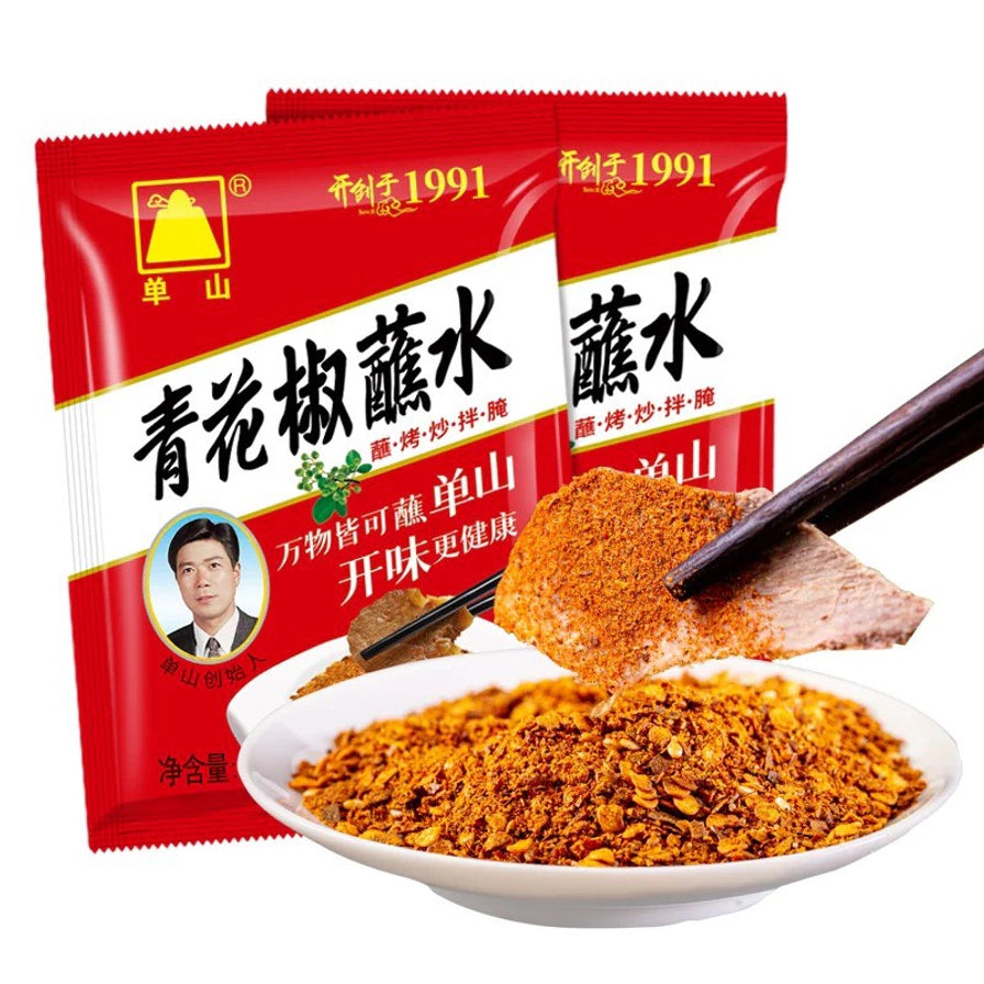 两袋单山青花椒蘸水包装，前方摆放一碟细腻的辣椒面，筷子夹着蘸满调料的肉片。
