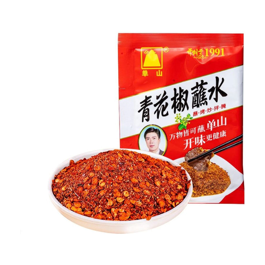 单山青花椒蘸水红色包装袋，前方摆放一碟装满红色辣椒粉和青花椒颗粒的干碟蘸料，背景纯白。