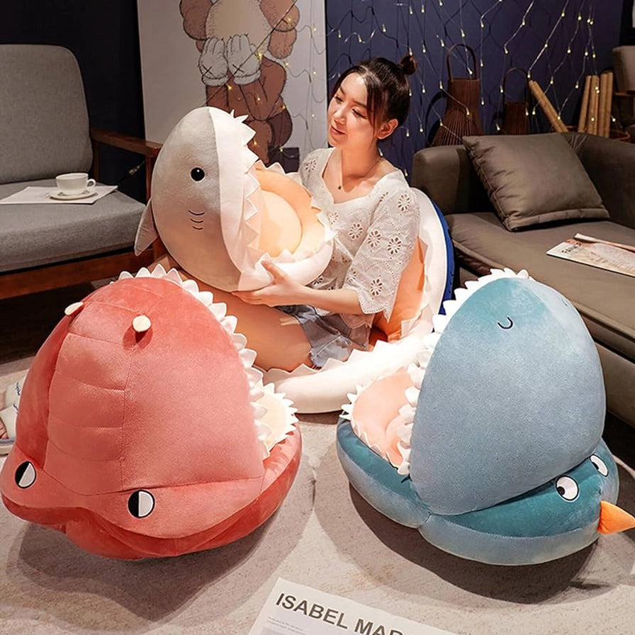 展示了粉红色、灰色和绿色三种不同款式的恐龙与鲨鱼造型坐垫，一名女性正抱着其中一款展示其柔软度。