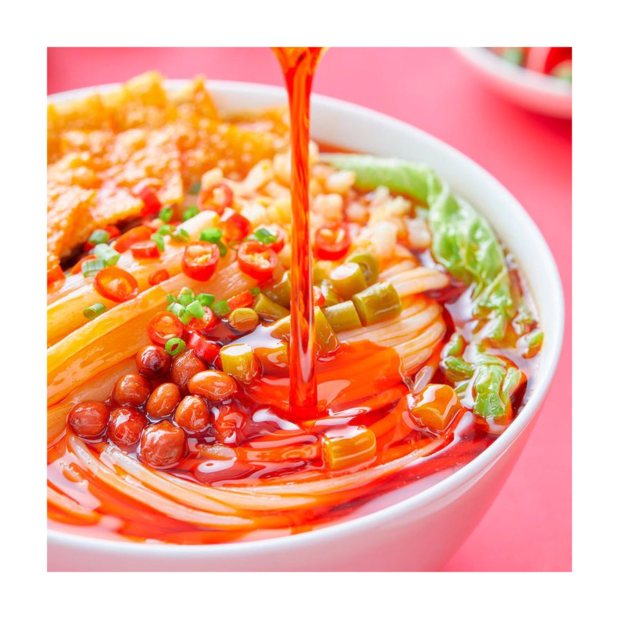 正在向螺蛳粉中倒入红亮辣油的近景特写，碗中可见米粉、花生、辣椒和酸豆角。