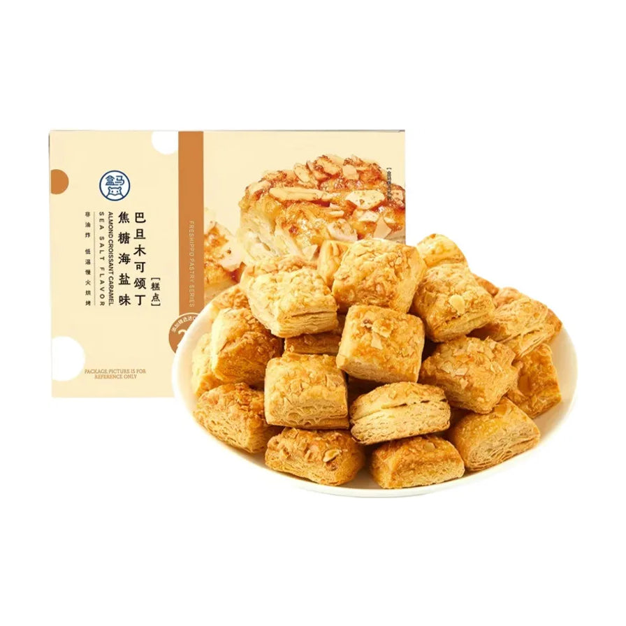 盒马焦糖海盐杏仁可颂方块，白色圆盘中堆叠着金黄酥脆的可颂方块，背景为产品包装盒，展示了巴旦木碎和层叠的酥皮纹理。