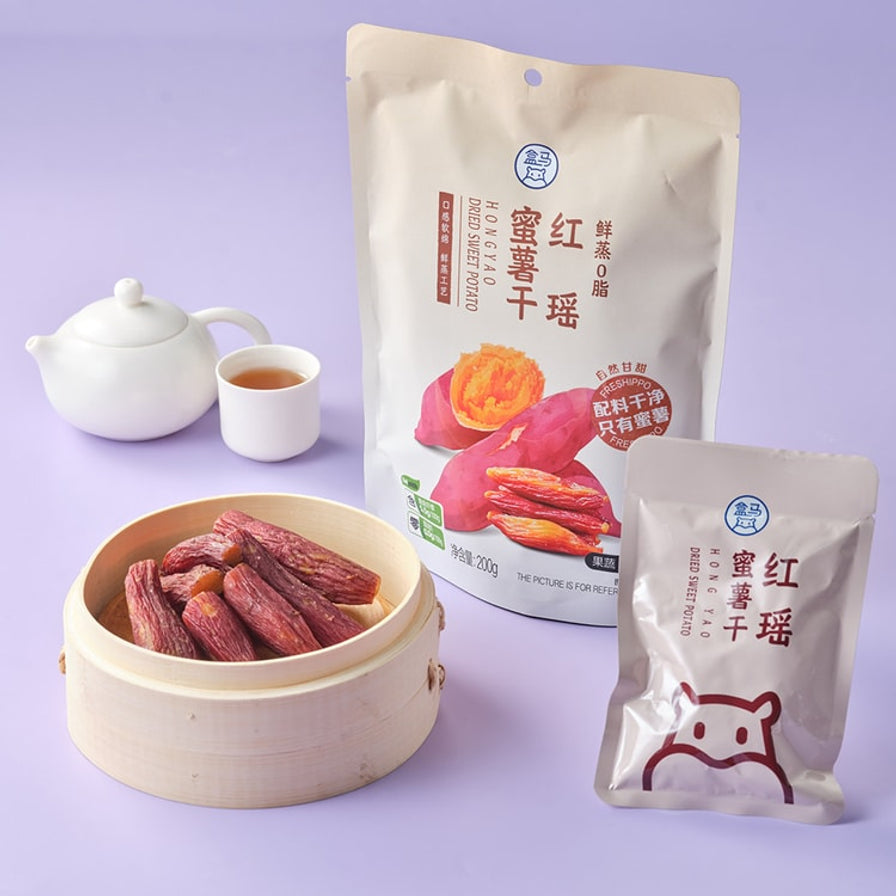 盒马鲜蒸0脂红瑶蜜薯干的大包装袋、独立小包装以及蒸笼中的红薯干，背景有茶壶茶杯。