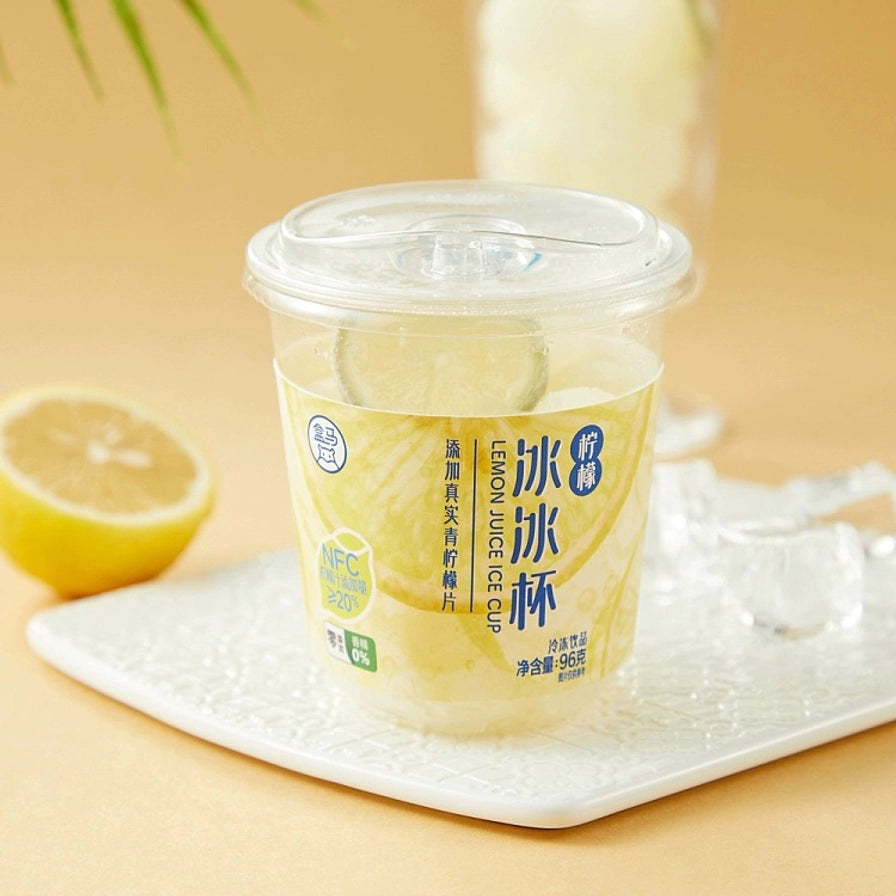 盒马柠檬冰冰杯在暖色背景下的生活化展示，杯子放置在白色托盘上，旁边配有新鲜柠檬和散落的冰块。