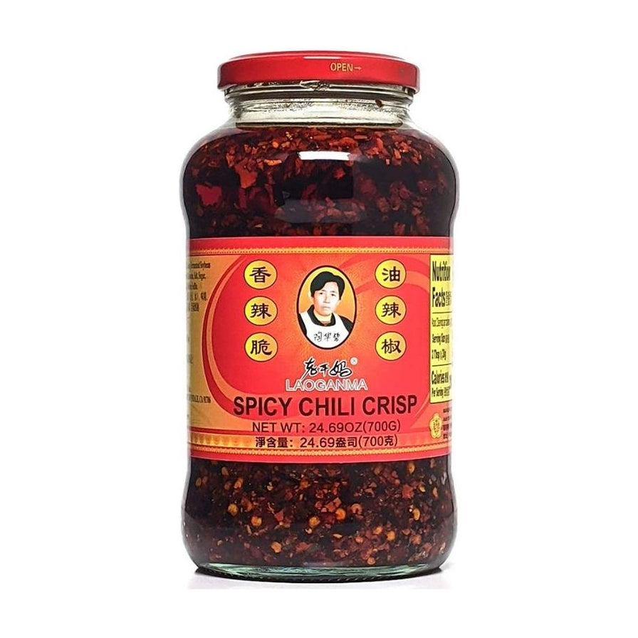 老干妈香辣脆油辣椒700克大瓶装正面图，透明玻璃瓶身可见内部红油与辣椒碎，标签印有陶华碧头像及Spicy Chili Crisp英文名。