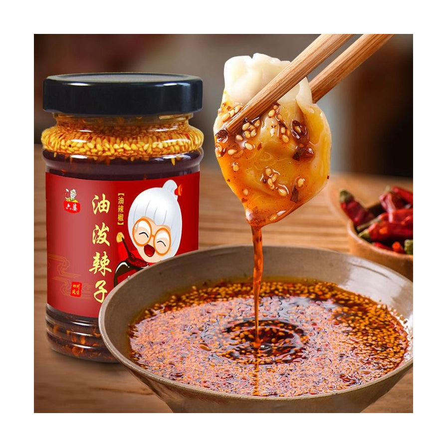 六婆油泼辣子展示，筷子夹起一只蘸满红亮辣椒油和芝麻的水饺，背景为产品包装瓶。