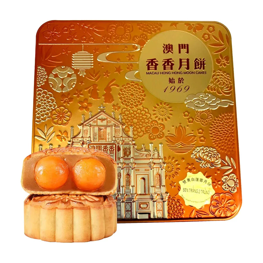 澳门香香双黄白莲蓉月饼礼盒，金色浮雕铁盒包装，前景展示切开的月饼，露出两颗饱满的咸蛋黄。