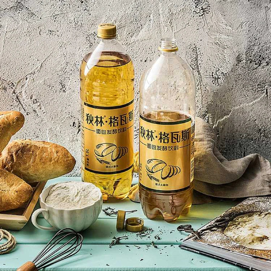两瓶秋林格瓦斯饮料摆放在桌面上，背景配有面包、面粉和搅拌器，展示其面包发酵的传统工艺背景。