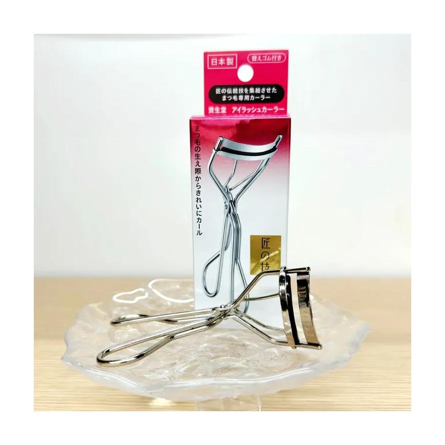 资生堂睫毛夹实物放置在透明玻璃托盘上，背景为产品包装盒，展示金属质感细节。