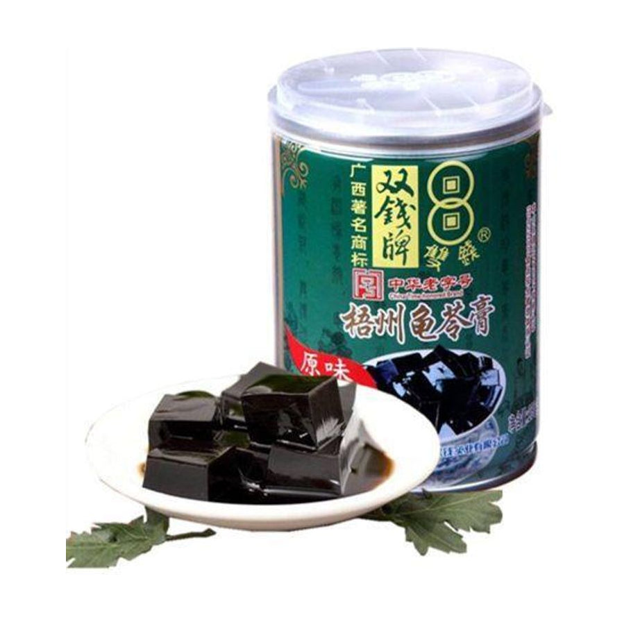 双钱牌梧州龟苓膏250g罐装，前景为白瓷碟盛放的Q弹黑色龟苓膏块，背景为绿色罐身包装。