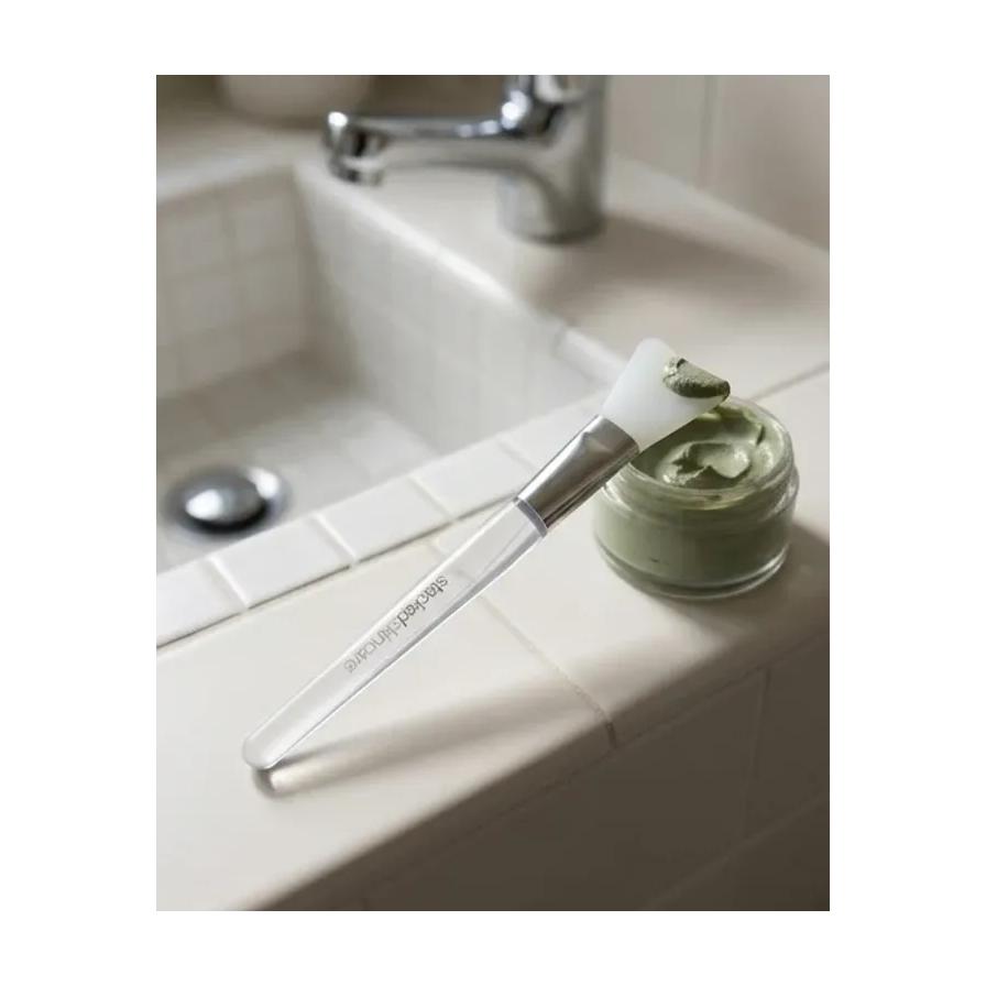 面膜刷置于浴室洗手台旁，刷头沾有绿色泥膜，旁边摆放着面膜罐的生活场景图。
