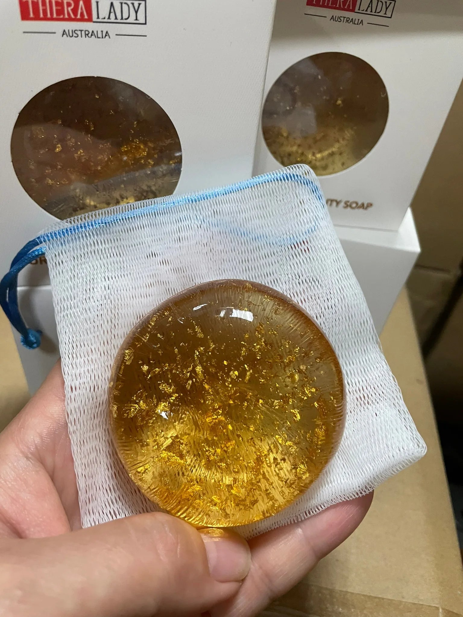 手持 Thera Lady 金箔美容皂特写，搭配白色起泡网袋，背景可见多盒库存包装。
