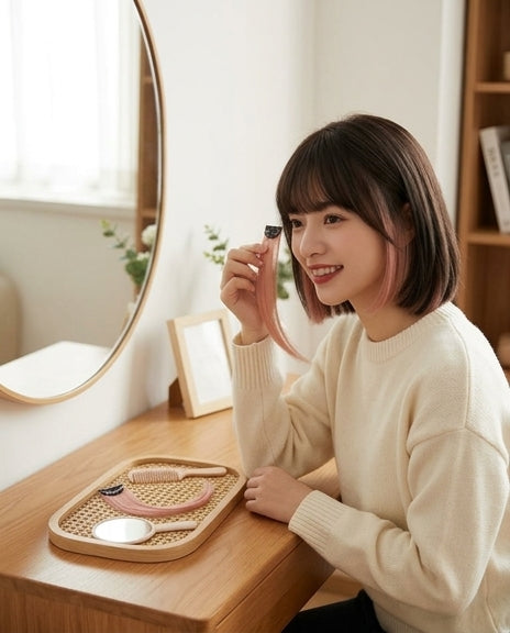 年轻女性坐在梳妆台前手持西柚粉假发片比对发型，桌面摆放梳子与镜子。