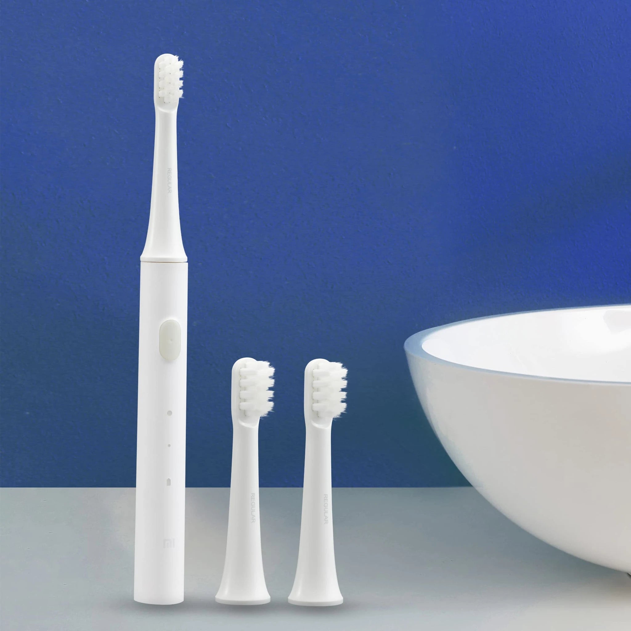 蓝色背景下的小米电动牙刷主机与两个替换刷头特写，旁边可见白色洗手盆边缘。
