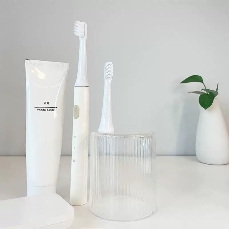 白色小米电动牙刷主机、替换刷头与透明收纳杯的桌面静物展示，背景有牙膏和绿植。