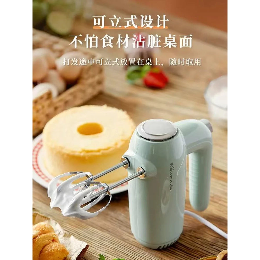 小熊手持电动打蛋器垂直站立在木质桌面上，背景有戚风蛋糕和烘焙食材，上方配有“可立式设计”中文说明。