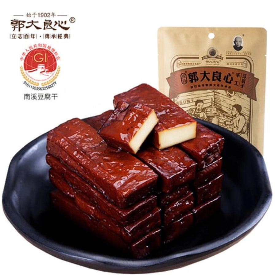 黑色圆盘中整齐叠放的深褐色手工豆腐干，质地油亮紧实，背景为产品包装袋及南溪豆腐干地理标志。