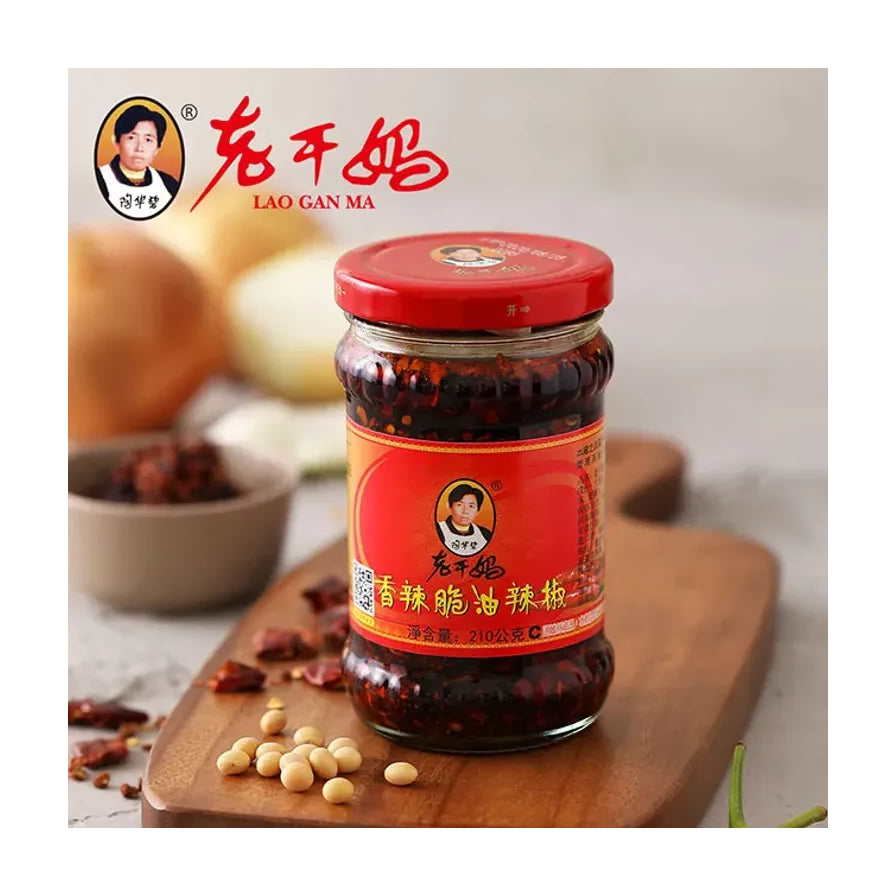 老干妈香辣脆油辣椒210克瓶装，放置在木质托盘上，背景配有大豆、干辣椒等食材，左上角带有老干妈品牌标志。