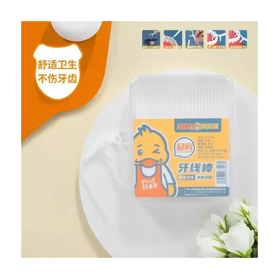 MEET DUCK 小黄鸭牙线棒生活场景图，盒装牙线棒放置在白色圆盘上，背景点缀有白色花朵，上方展示了产品使用细节。