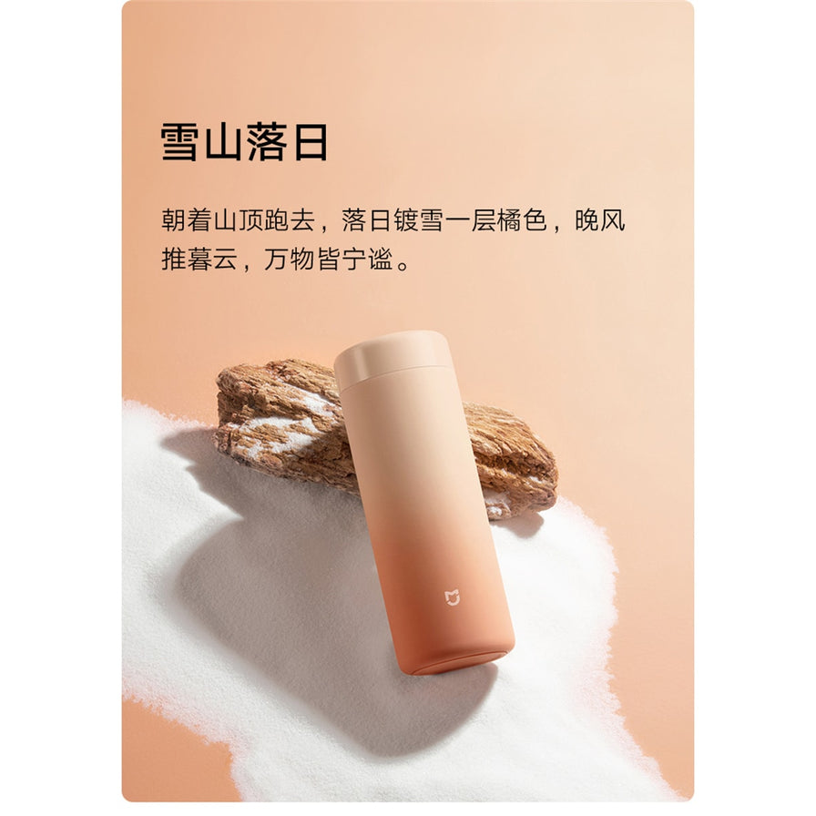 米家渐变色保温杯斜放在白色沙粒和木块背景上，配有雪山落日主题文字描述。