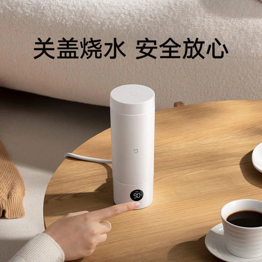 小米电热杯放置在圆桌上，手指正在触碰底部的温度显示屏，显示90度，上方文字为“关盖烧水 安全放心”。