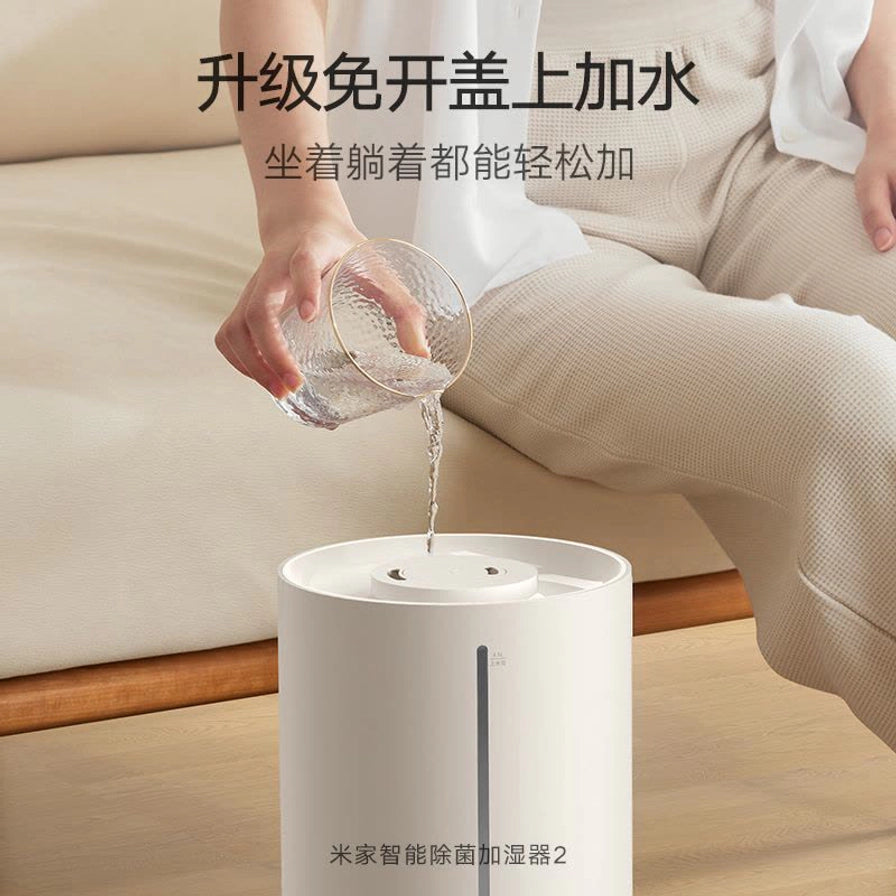 展示加湿器免开盖上加水功能，一人正将水杯中的水直接倒入加湿器顶部，背景为居家沙发环境。